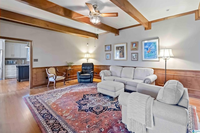 living area with ceiling fan, beamed ceiling, light wood-style flooring, and wainscoting