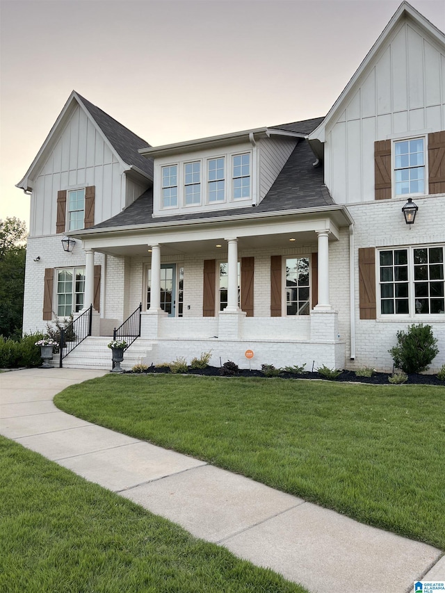 modern farmhouse with a front yard, covered porch, a shingled roof, board and batten siding, and brick siding