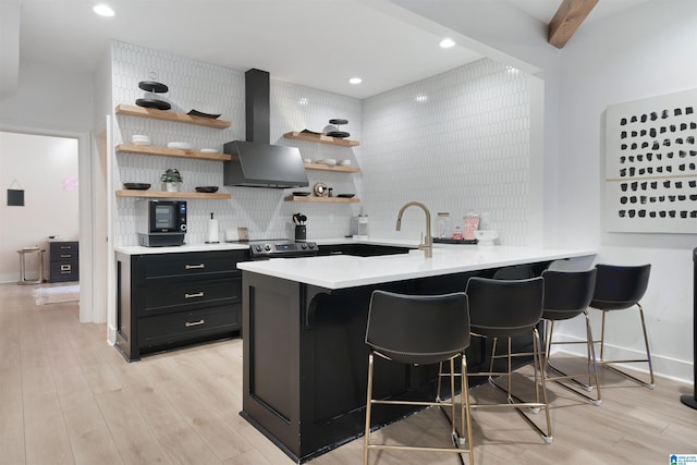 kitchen with backsplash, open shelves, a breakfast bar area, wall chimney exhaust hood, and dark cabinets