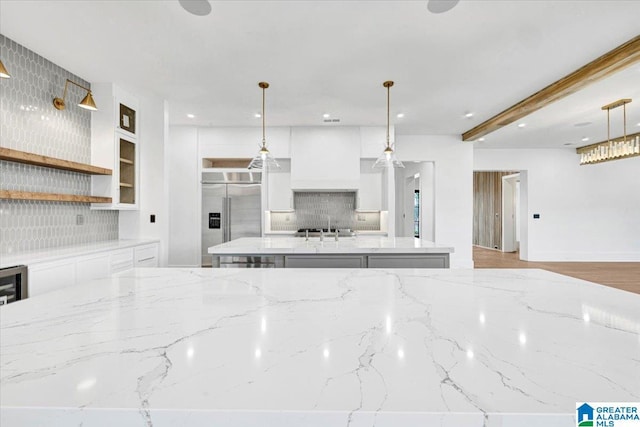 kitchen featuring premium range hood, a kitchen island with sink, white cabinets, built in fridge, and backsplash