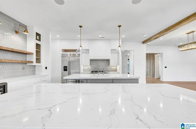 kitchen featuring backsplash, light stone counters, built in fridge, white cabinetry, and a kitchen island with sink