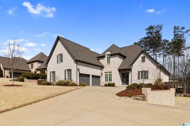 french country inspired facade featuring an attached garage, roof with shingles, concrete driveway, and brick siding