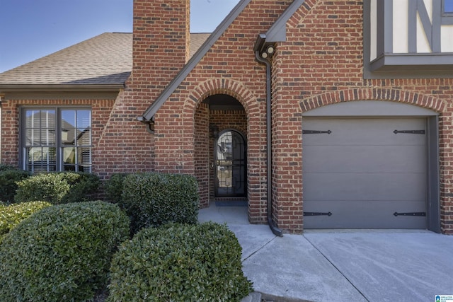 property entrance featuring a chimney, concrete driveway, brick siding, and a shingled roof