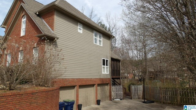 view of home's exterior with driveway, roof with shingles, an attached garage, fence, and brick siding