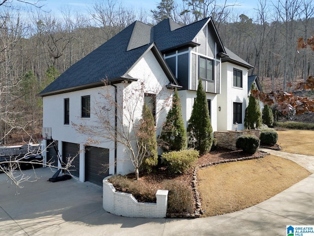 view of front of property featuring a garage, driveway, stone siding, a forest view, and roof with shingles