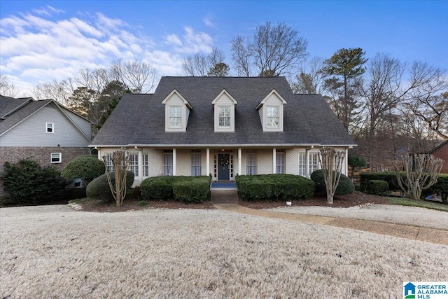 cape cod home featuring covered porch, brick siding, and roof with shingles