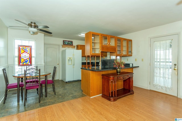 kitchen featuring white refrigerator with ice dispenser, wood finished floors, brown cabinets, dark countertops, and glass insert cabinets