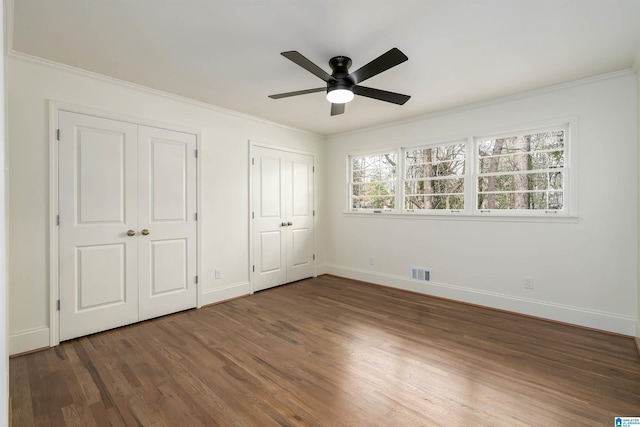 unfurnished bedroom featuring wood finished floors, visible vents, baseboards, ornamental molding, and multiple closets