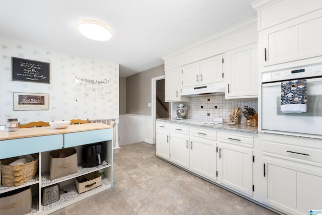 kitchen featuring a wainscoted wall, white cabinetry, light stone countertops, oven, and under cabinet range hood