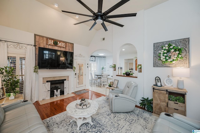 living area featuring wood finished floors, a ceiling fan, high vaulted ceiling, a fireplace, and arched walkways