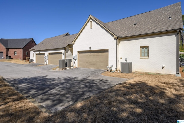 view of side of property featuring brick siding, central AC unit, a garage, and roof with shingles