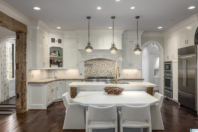 kitchen with a kitchen island with sink, a sink, white cabinetry, arched walkways, and built in appliances