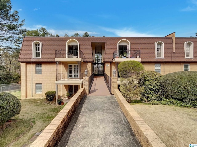 view of front facade with mansard roof, brick siding, a balcony, and fence