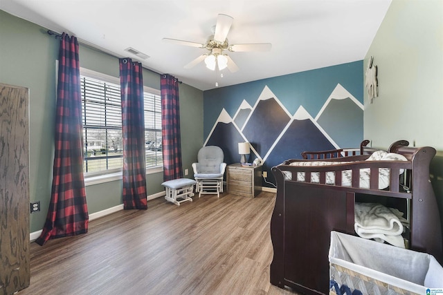 bedroom featuring visible vents, an accent wall, ceiling fan, baseboards, and wood finished floors