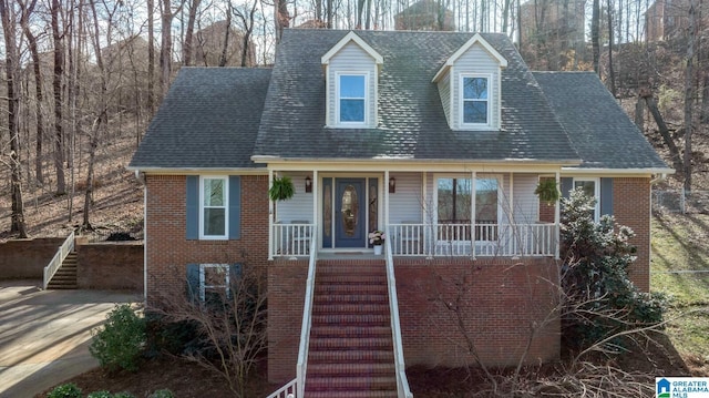 new england style home with brick siding, a porch, a shingled roof, and stairs