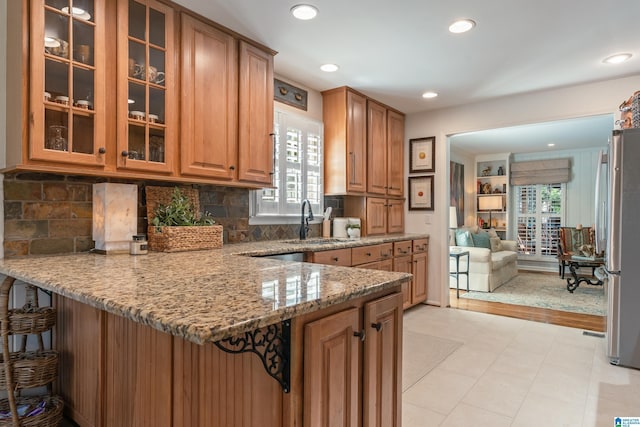 kitchen with a sink, freestanding refrigerator, a peninsula, glass insert cabinets, and light stone countertops