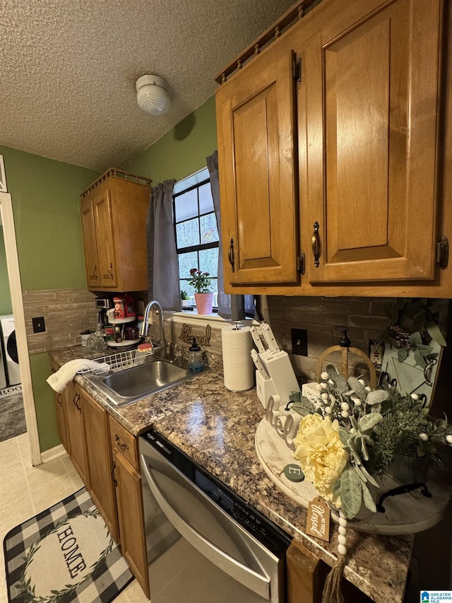 kitchen featuring washer / dryer, stainless steel dishwasher, brown cabinetry, a textured ceiling, and a sink