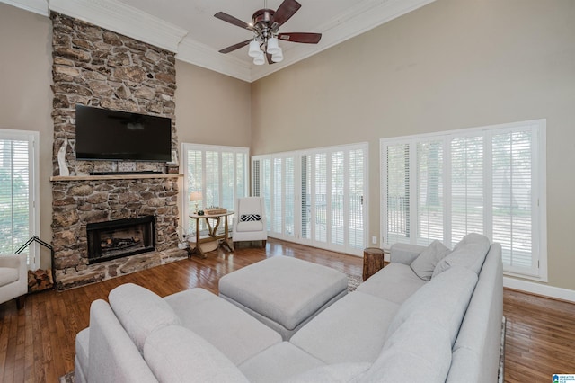 living area with a ceiling fan, wood finished floors, ornamental molding, a stone fireplace, and a towering ceiling