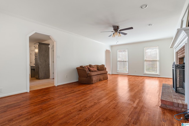 unfurnished living room with a brick fireplace, crown molding, light wood-style flooring, arched walkways, and a ceiling fan