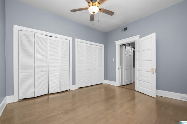 unfurnished bedroom featuring a ceiling fan, visible vents, wood finished floors, baseboards, and multiple closets