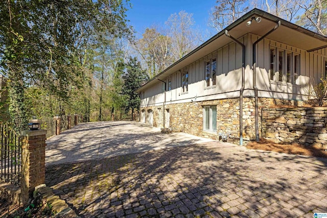 view of property exterior featuring stone siding, board and batten siding, decorative driveway, and fence