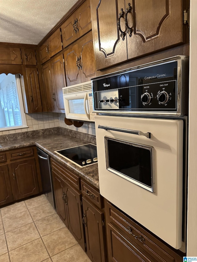 kitchen featuring backsplash, dark countertops, a textured ceiling, white appliances, and light tile patterned floors