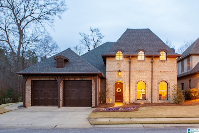 french country inspired facade with a garage, brick siding, concrete driveway, and a shingled roof