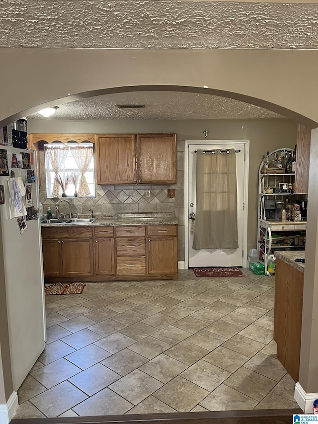 kitchen with brown cabinetry, arched walkways, freestanding refrigerator, a sink, and backsplash