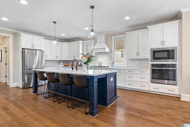 kitchen featuring premium range hood, an island with sink, stainless steel appliances, light countertops, and white cabinets