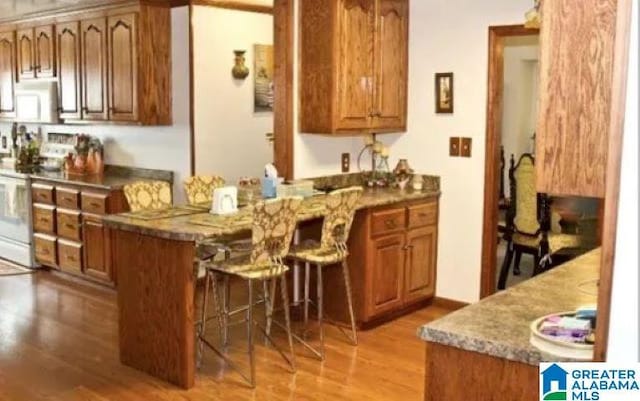 kitchen with brown cabinets, wood finished floors, white electric range oven, a peninsula, and a breakfast bar area