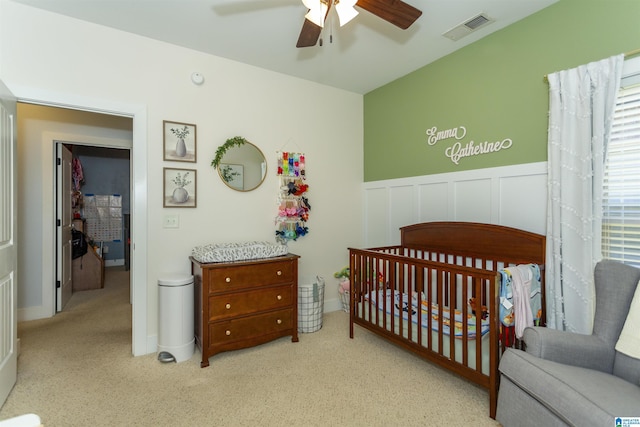 carpeted bedroom featuring visible vents, a crib, wainscoting, a decorative wall, and a ceiling fan