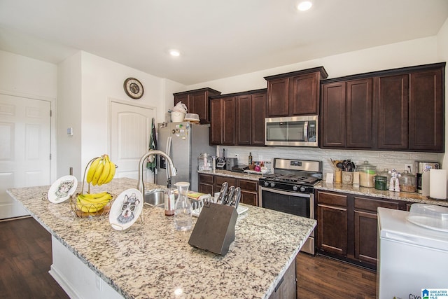 kitchen featuring backsplash, dark brown cabinets, dark wood finished floors, a center island with sink, and appliances with stainless steel finishes