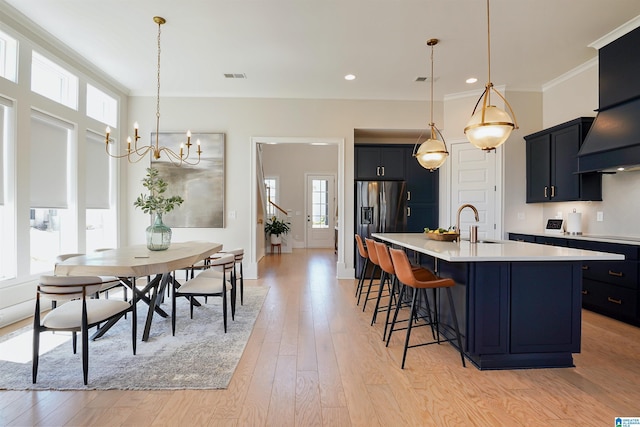 kitchen with stainless steel fridge with ice dispenser, a chandelier, a center island with sink, light wood-type flooring, and light countertops