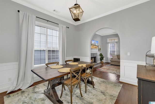 dining space featuring visible vents, a wainscoted wall, a warm lit fireplace, arched walkways, and wood-type flooring