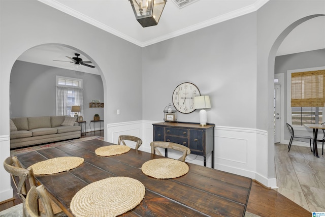 dining area with a wainscoted wall, crown molding, a ceiling fan, and arched walkways