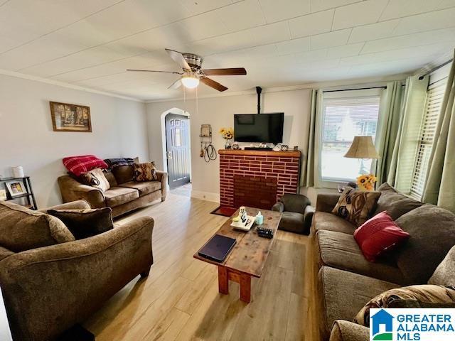 living area with crown molding, a brick fireplace, a ceiling fan, and light wood-type flooring