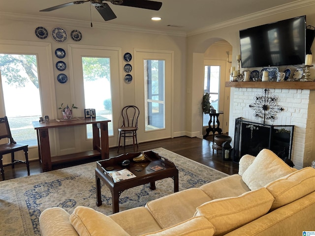 living area featuring wood finished floors, a ceiling fan, arched walkways, crown molding, and a brick fireplace