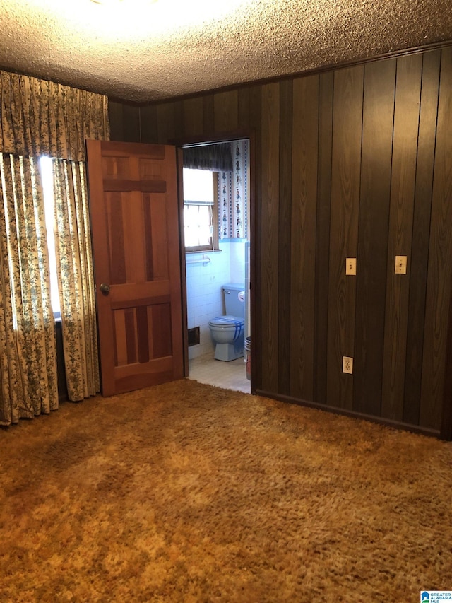 unfurnished bedroom with carpet floors, wood walls, ensuite bath, and a textured ceiling