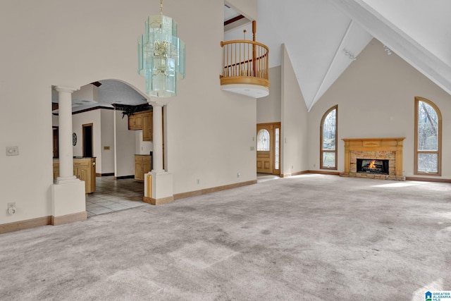 unfurnished living room with high vaulted ceiling, a brick fireplace, ornate columns, baseboards, and light colored carpet