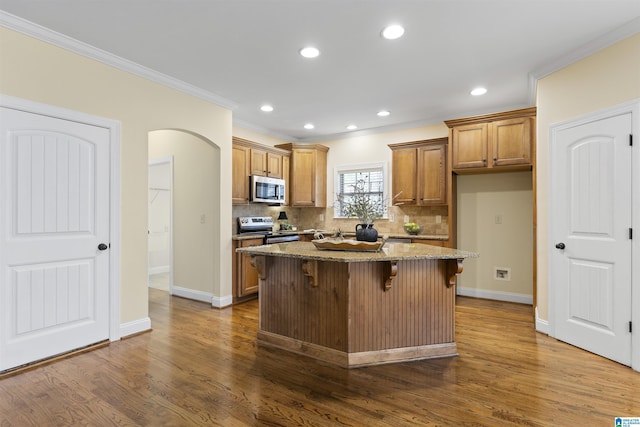 kitchen featuring light stone counters, appliances with stainless steel finishes, brown cabinets, ornamental molding, and dark wood-style flooring