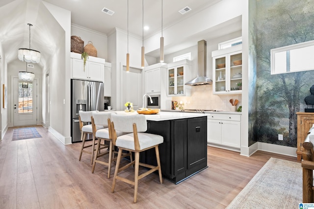 kitchen with light wood finished floors, visible vents, wall chimney range hood, a kitchen island with sink, and appliances with stainless steel finishes