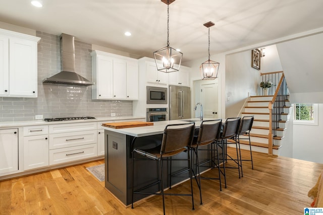 kitchen featuring light wood-style floors, an island with sink, wall chimney range hood, appliances with stainless steel finishes, and light countertops
