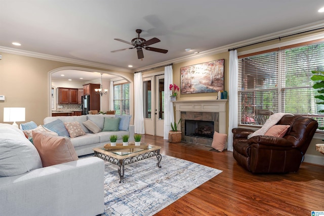 living room with arched walkways, a tiled fireplace, ornamental molding, wood finished floors, and ceiling fan with notable chandelier