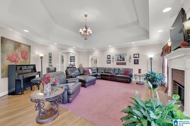 living area with an inviting chandelier, a brick fireplace, a tray ceiling, arched walkways, and wood finished floors