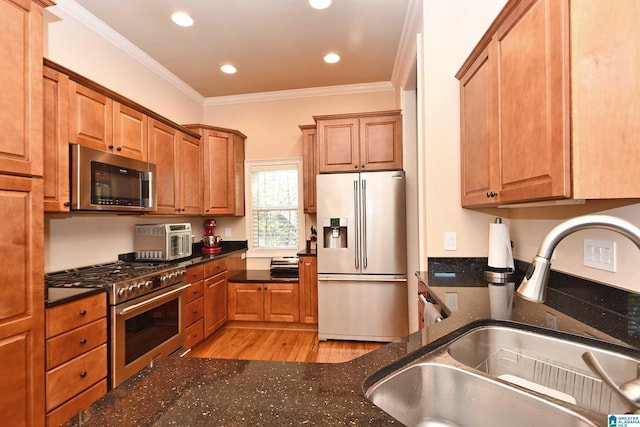 kitchen with stainless steel appliances, a sink, light wood-style floors, crown molding, and dark stone counters