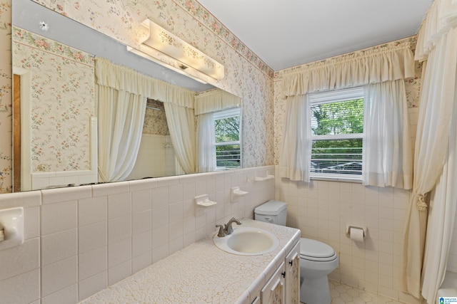 full bathroom with tile walls, toilet, vanity, wallpapered walls, and a wainscoted wall