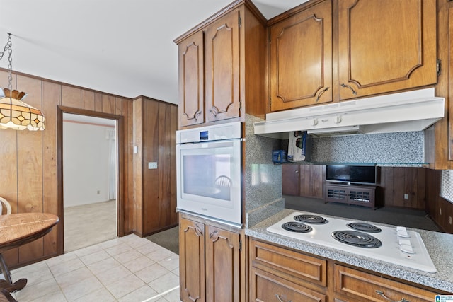 kitchen featuring white appliances, under cabinet range hood, wood walls, light countertops, and brown cabinets