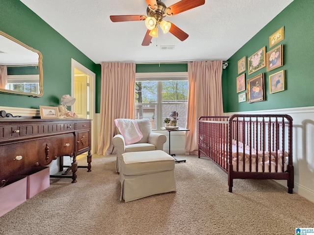 carpeted bedroom with a textured ceiling, visible vents, a crib, and a ceiling fan