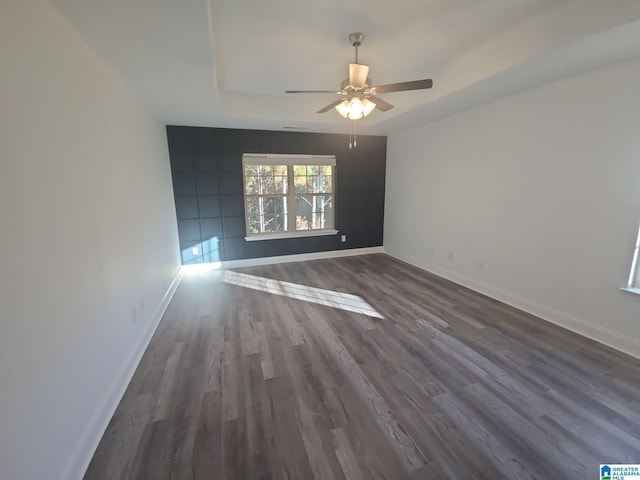 unfurnished room with a tray ceiling, ceiling fan, baseboards, and dark wood-style floors