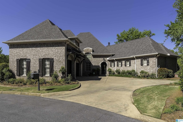 french country inspired facade featuring brick siding, curved driveway, and roof with shingles
